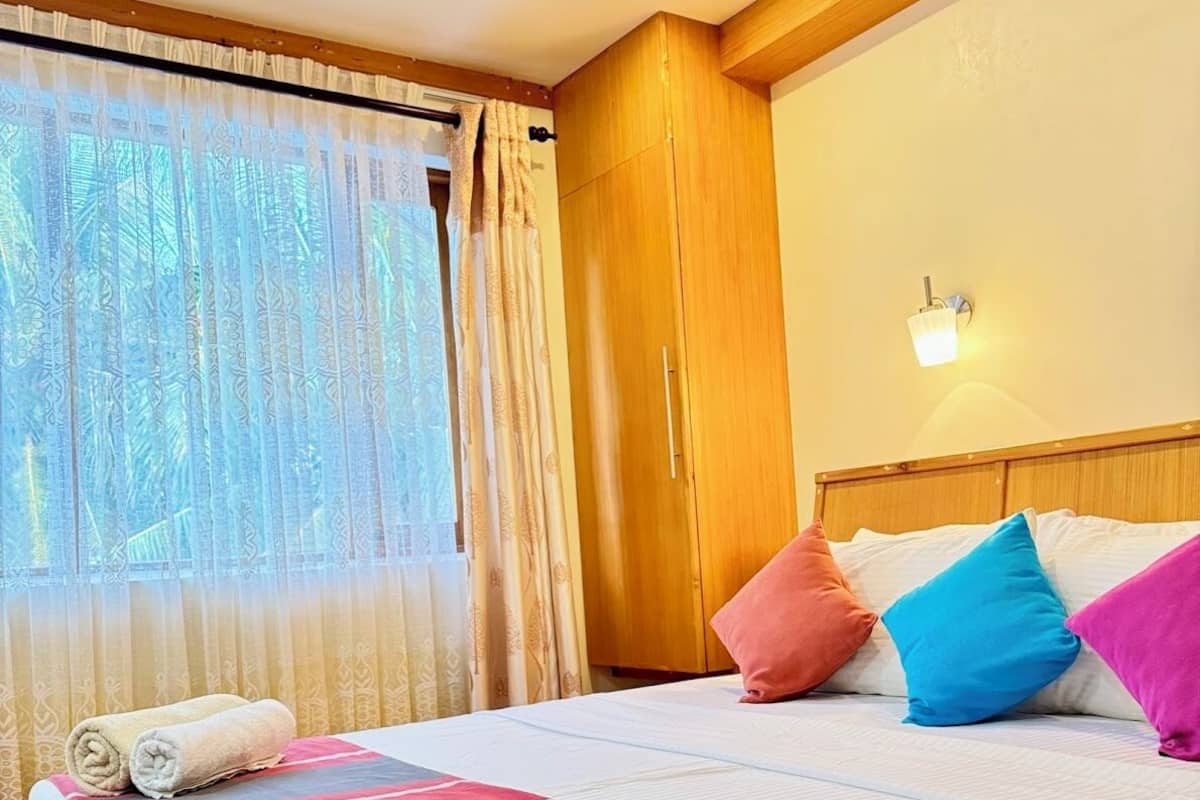 Une chambre d’hôtel avec un lit, des oreillers, une fenêtre avec des rideaux et une lampe fixée au mur.