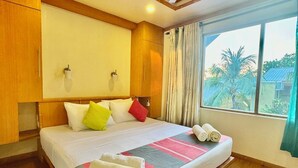 A hotel room with a large bed, wooden headboard, two wall-mounted lights, a window with curtains, and a view of palm trees outside.