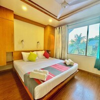 Habitación de hotel con una cama grande, cabecero de madera, dos luces fijadas a la pared, una ventana con cortinas y vistas a palmeras fuera.