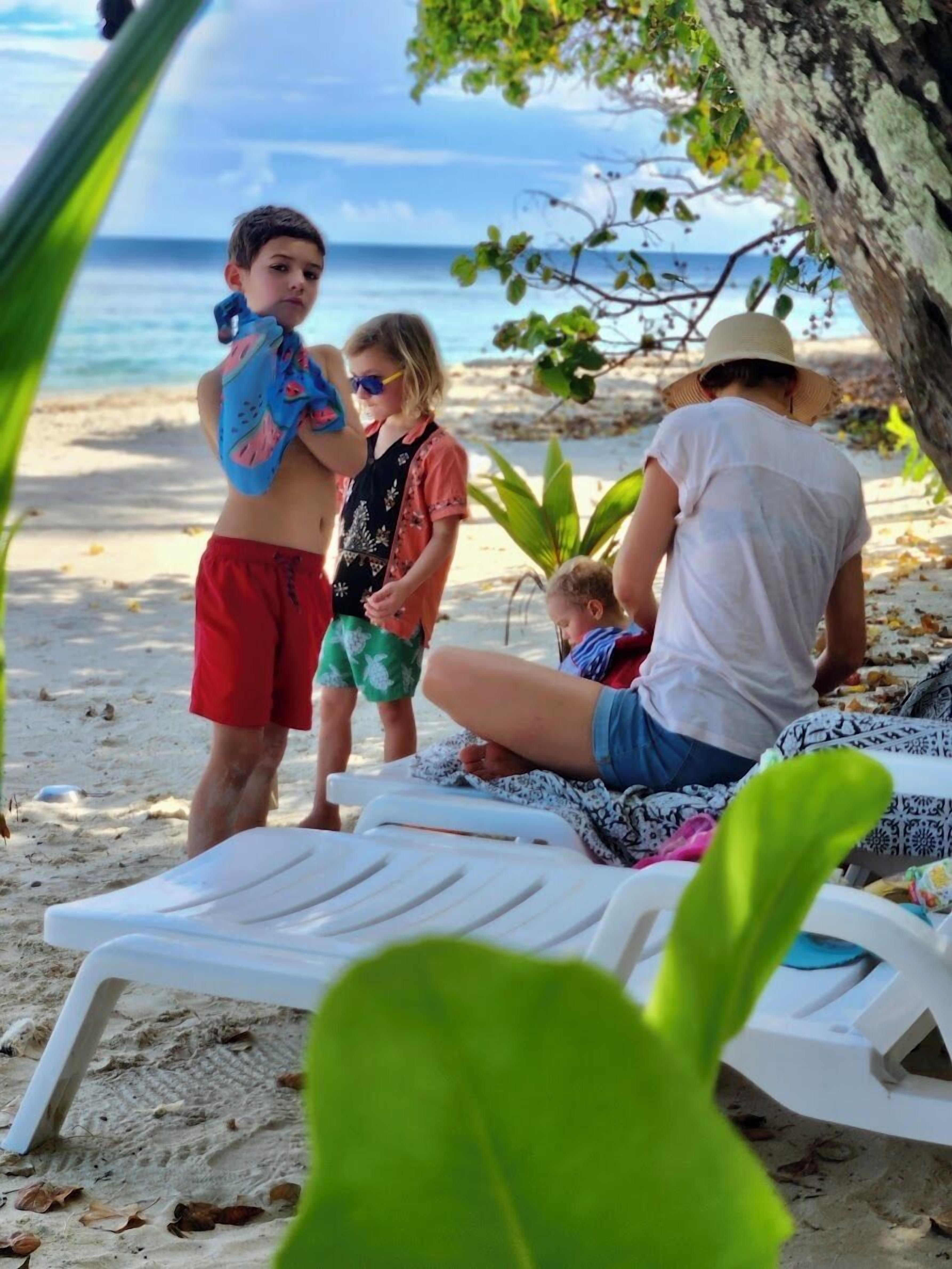 Eine Strandansicht mit einer Familie im Sand, einer weißen Liege und einem Baum.
