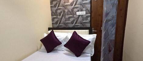 A single bed with white bedding and two purple pillows, a geometric-patterned wall, and a wooden door.