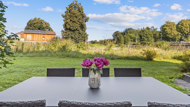 Una mesa de comedor con un jarrón de flores, rodeada de sillas de mimbre, ubicada en un espacio al aire libre con césped y vistas a un edificio y árboles.