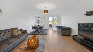 Una sala moderna con un sofá marrón oscuro, una mesa de centro y un televisor. Se observa un comedor con una mesa y sillas al fondo.