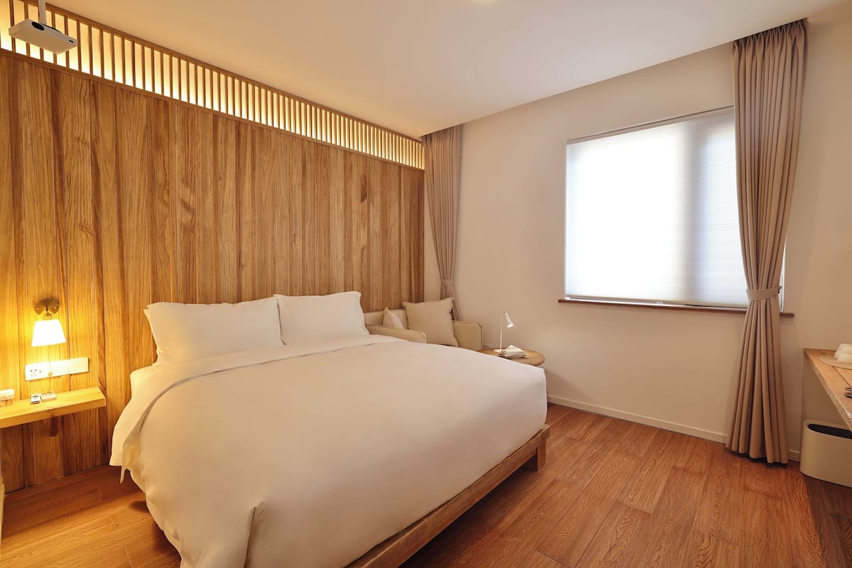 Une chambre d’hôtel avec une tête de lit en bois, un grand lit, une table de chevet avec une lampe, une commode et une fenêtre avec des rideaux.