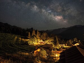 Een nachtelijke scène van een bergachtig gebied met verschillende verlichte tenten en hutjes, voor een sterrenhemel.