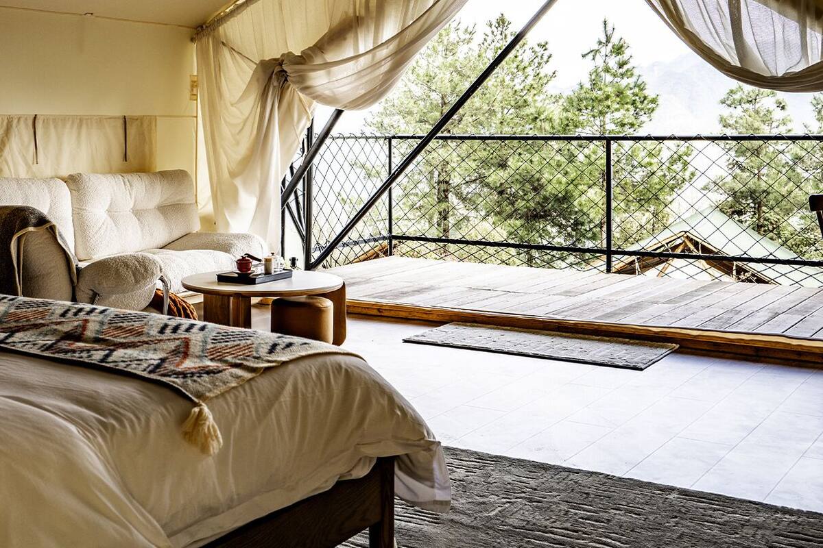 A bedroom with a large bed, a bedside table with books, a chair, and a balcony with a view of trees.