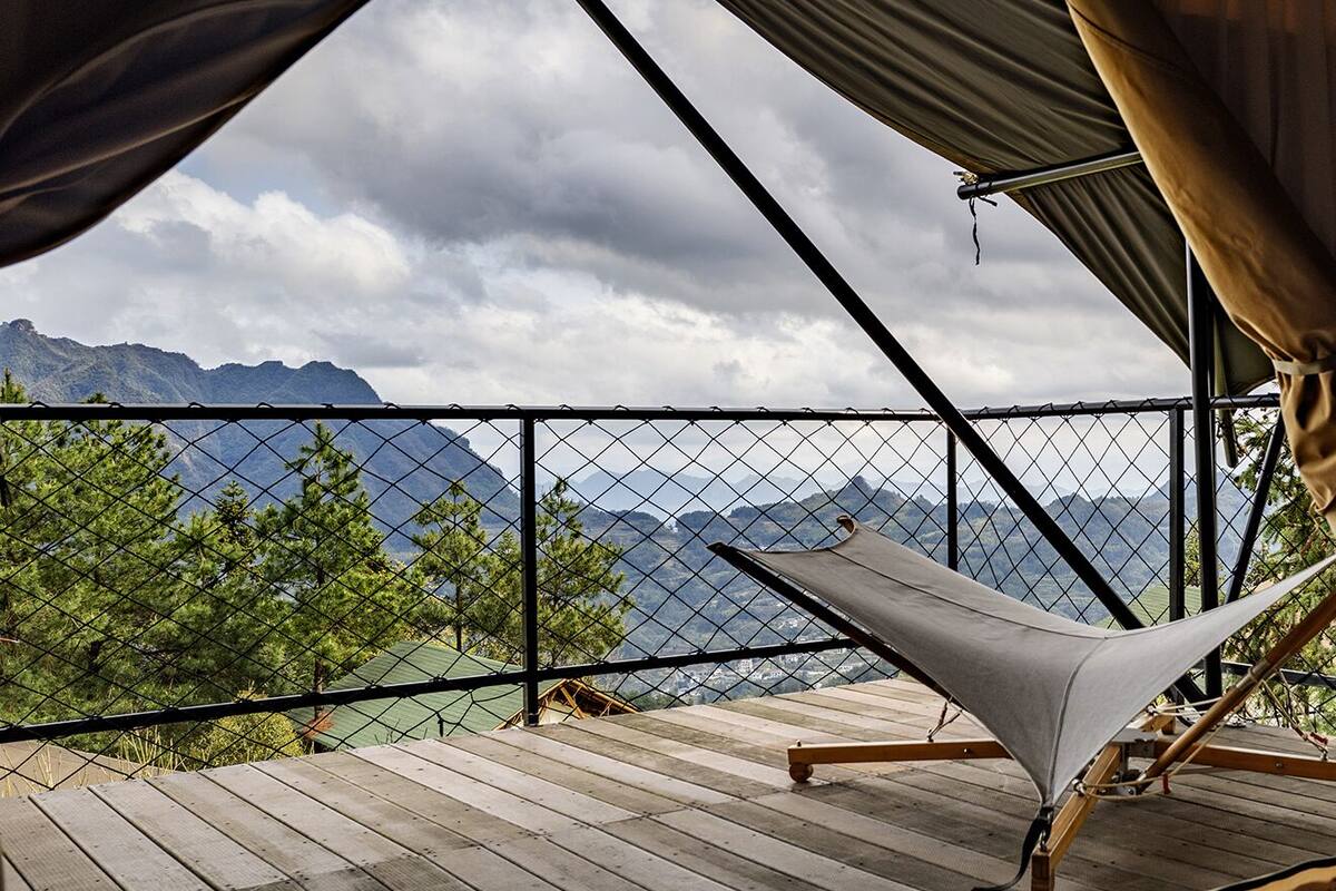 A hammock on a wooden deck with a view of mountains and trees through a chain-link fence.