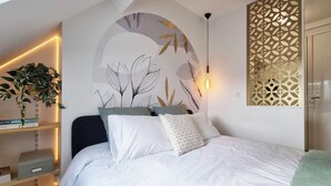 A bedroom with a bed, pillows, a wall-mounted light, a decorative plant, and a bookshelf.