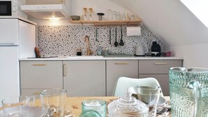 A modern kitchen with white cabinets, a white countertop, and a backsplash with a mosaic pattern. There are various kitchen appliances, including a microwave, oven, and coffee maker. A wooden shelf above the sink holds glassware and a tea kettle.