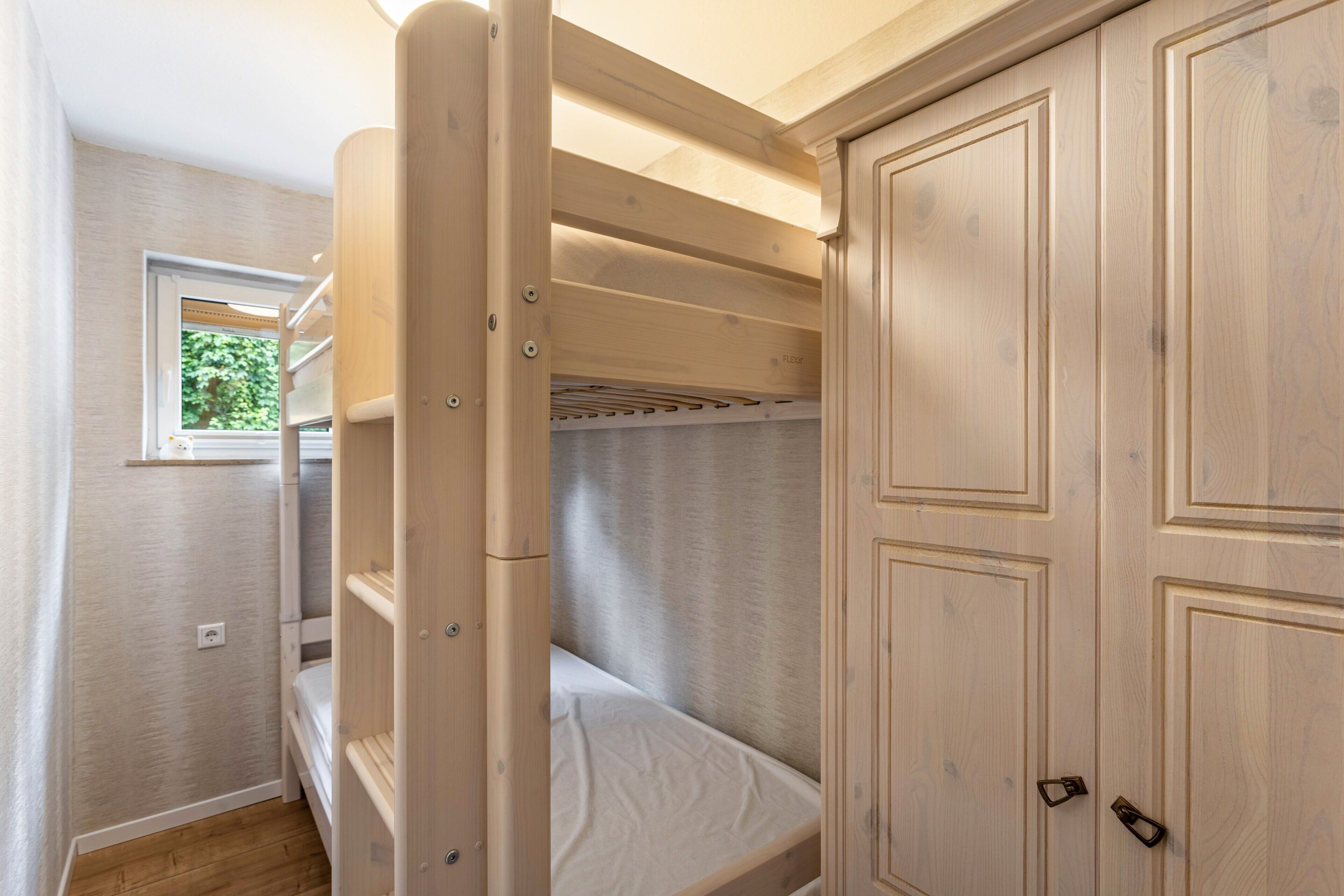 A bunk bed with a window, wooden wardrobe, and a light switch on the wall.