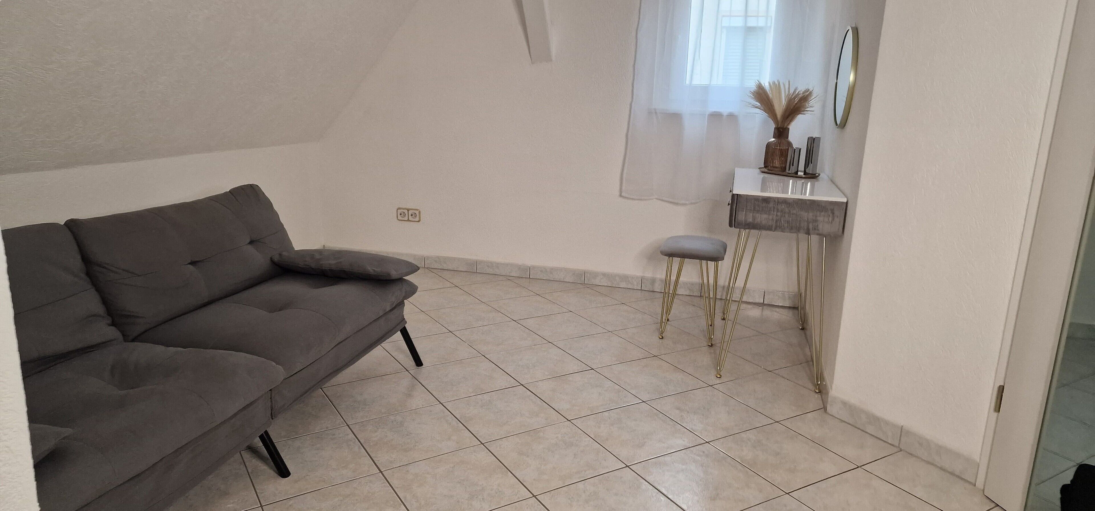 Ein Zimmer mit grauem Sofa, einem kleinen Hocker, einem weißen Konsolentisch mit Dekoration und einem Fenster mit transparenten Vorhängen.