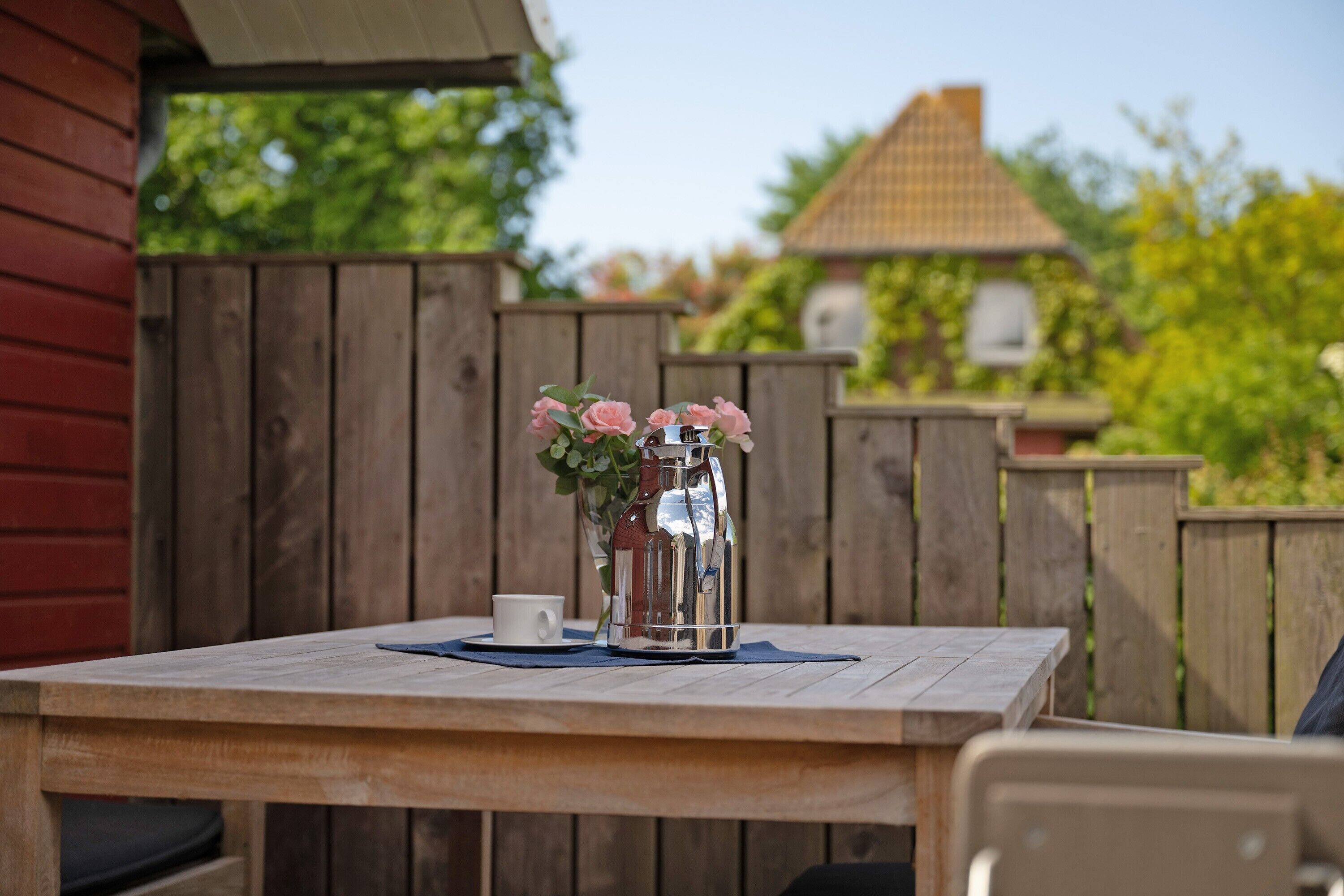 A wooden table with a metal pitcher and a cup, surrounded by a wooden fence and greenery.