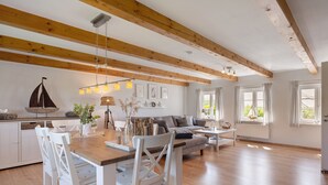 A spacious living area with a wooden ceiling, a dining table with chairs, a sofa, and a kitchen area with white cabinets. - Holiday Home 'Ferienhaus Pellworm Findling' with Terrace,  Garden and Wi-Fi (Pellworm)