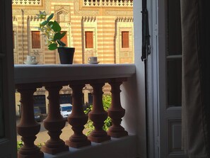 Ein Balkon mit einem Blumentopf, zwei Tassen und Blick auf ein Gebäude mit gemusterten Fenstern.