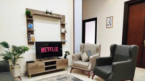 Ett vardagsrum med en platt-TV som visar "NETFLIX", en beige fåtölj, en mörk fåtölj, en TV-möbel i trä och en mönstrad matta.