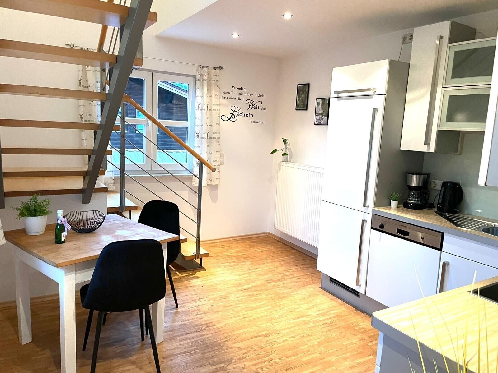 Une cuisine moderne avec des meubles blancs, un coin repas avec une table et des chaises en bois, un escalier et une fenêtre avec des rideaux.