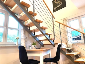Un interior moderno con una escalera de madera, un comedor con dos sillas y una mesa con una botella y un tazón.
