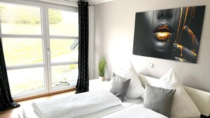 Ein Schlafzimmer mit Bett, Kissen, einem Nachttisch, einem Fenster mit Vorhängen und einem an der Wand hängenden Kunstwerk.