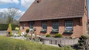 Une maison en briques avec un toit en pente, de nombreuses fenêtres et un jardin devant, agrémenté de diverses plantes et d’une clôture en bois.