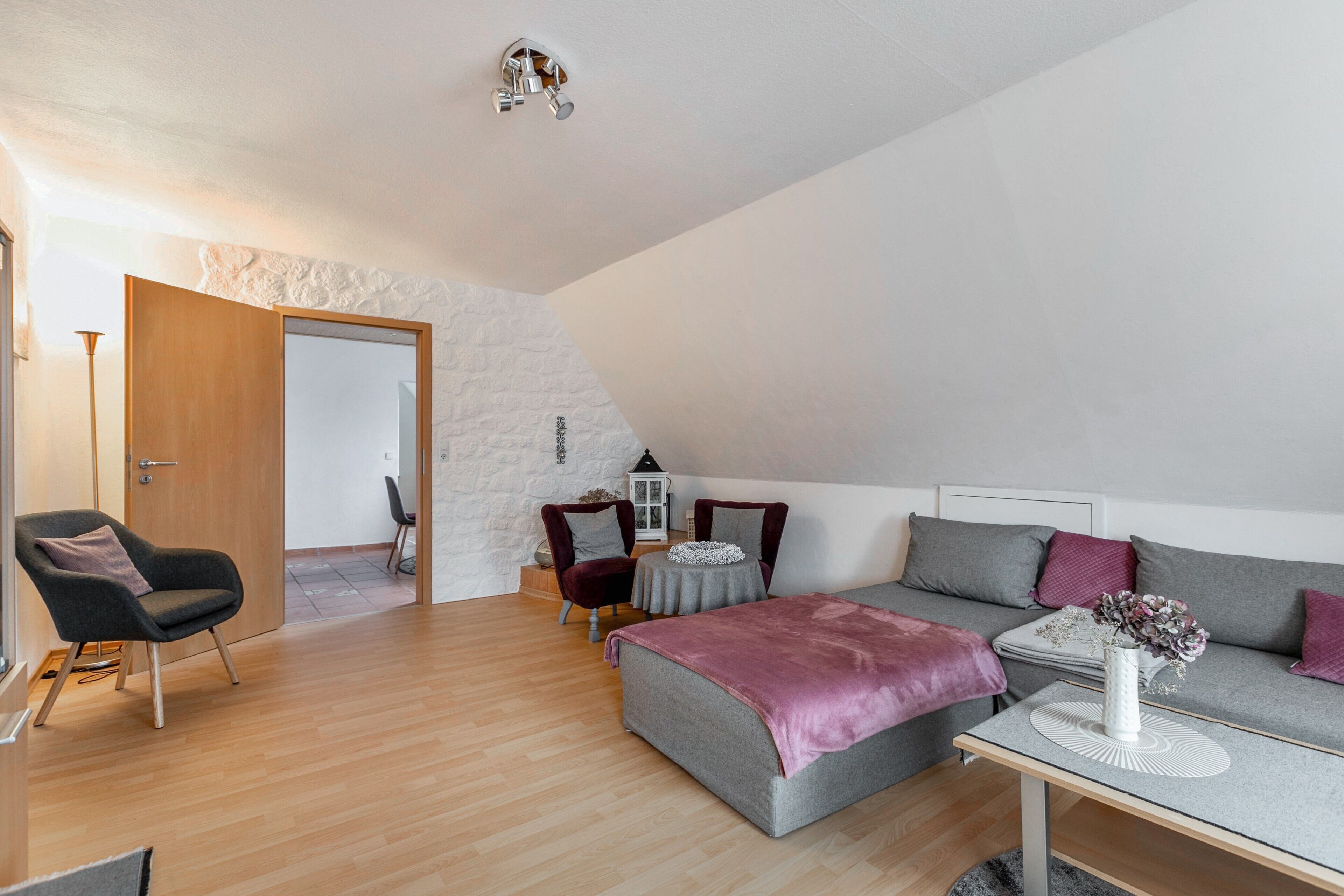 A modern loft bedroom with a sloped ceiling, a grey sofa, a small round table, and a chair.