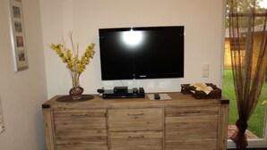 Un mueble de madera con un televisor de pantalla plana, un florero con flores amarillas y una mesita con controles remotos.
