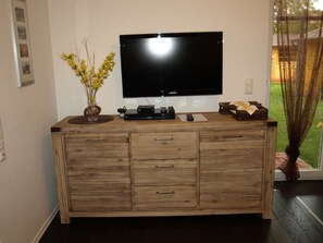 A wooden entertainment center with a flat-screen TV, a vase with yellow flowers, and a small table with remote controls.