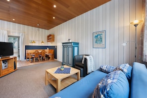 Une pièce comprenant un canapé bleu, une table basse en bois, un téléviseur posé sur un meuble en bois, un coin repas avec des chaises et une table, et un coin cuisine avec des placards et un appareil électroménager bleu.