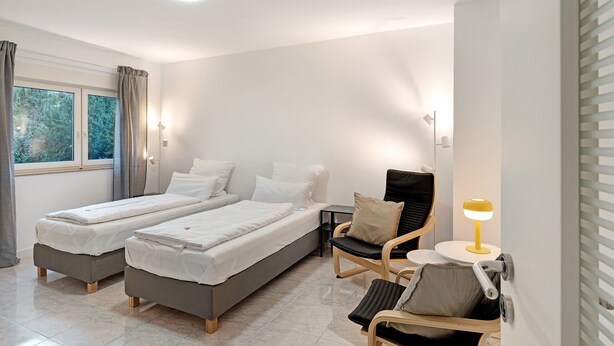 A modern bedroom with two beds, a chair, a small table, and a window with curtains. - Apartment 'Die Edisonstraße' with Wi-Fi (Pforzheim)