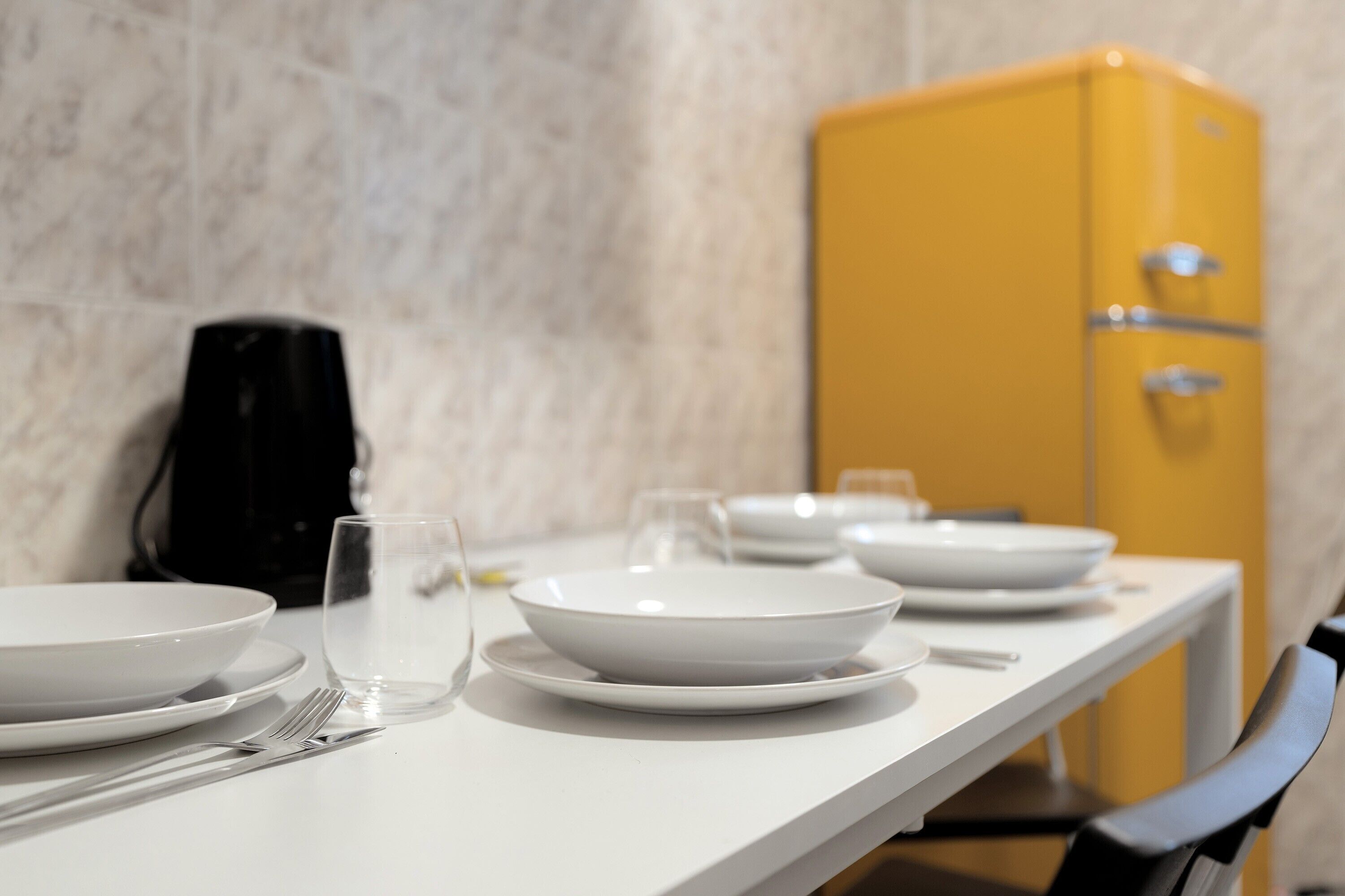 Une table à manger avec des assiettes blanches, une fourchette et un verre d’eau. Un buffet jaune se trouve à l’arrière-plan.