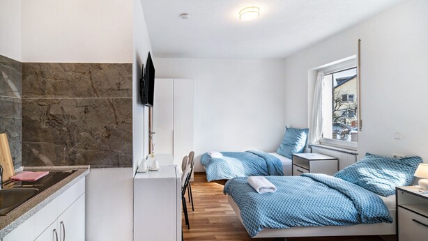 A modern hotel room with a kitchen area, two beds with blue bedding, a white wardrobe, and a window with a view. - Room 'Zimmer 4' with Wi-Fi (Berg-Weiler)
