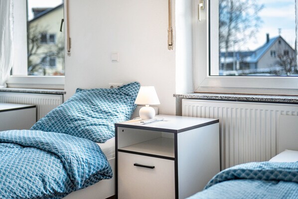 A bedroom with a bed, a nightstand with a lamp, and a window with a view of a house.