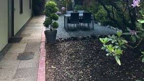 A covered outdoor area with a table and chairs, surrounded by greenery and purple flowers. - Apartment 'Das Spatzennest' with Sauna, Mountain Views & Private Terrace (Bad Harzburg)