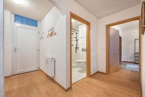 Un couloir avec un plancher en bois, une porte blanche, une salle de bain avec une douche et un radiateur.