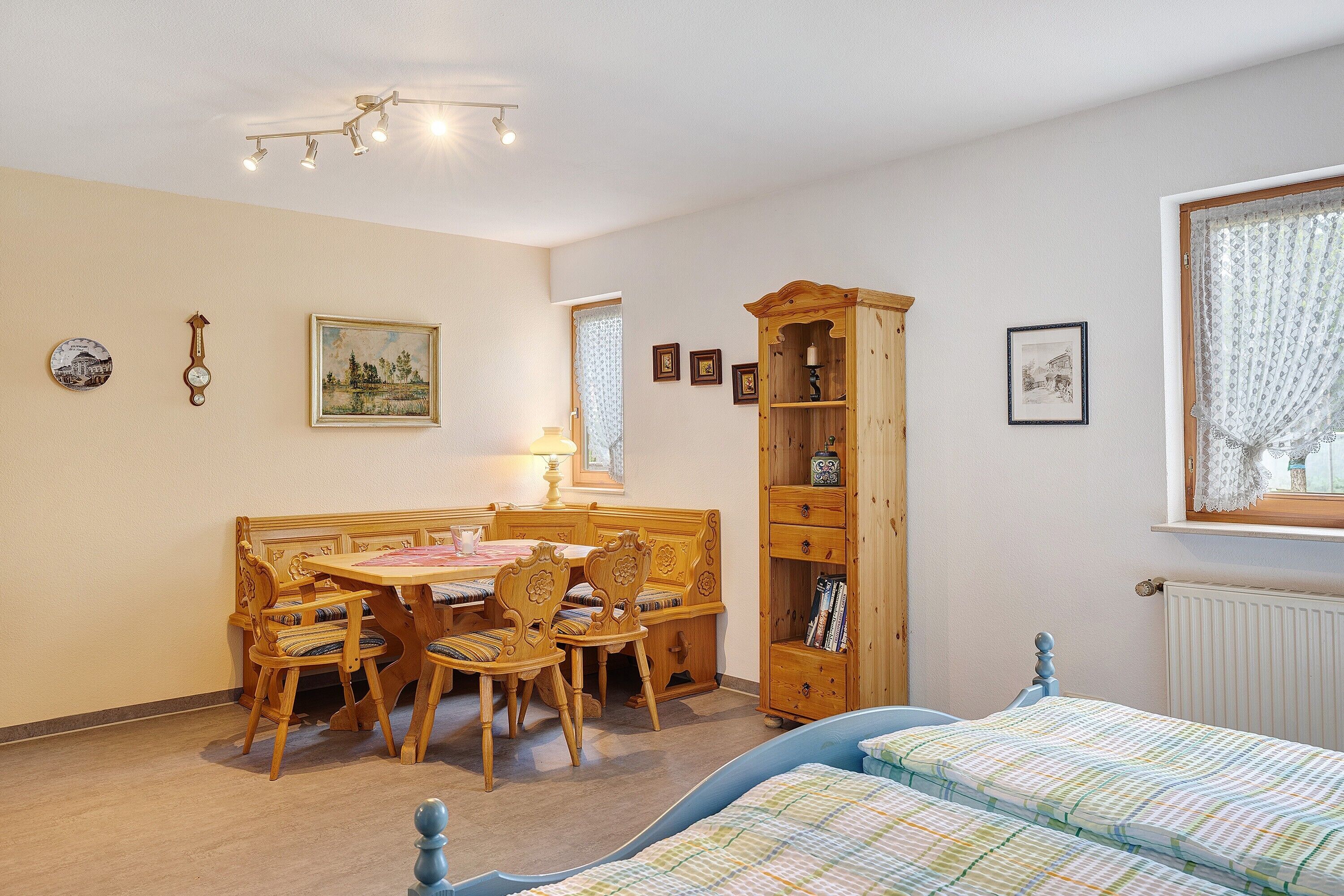 Una habitación con una mesa de comedor de madera, sillas, una estantería, una cama con colcha a rayas y una ventana con cortina.