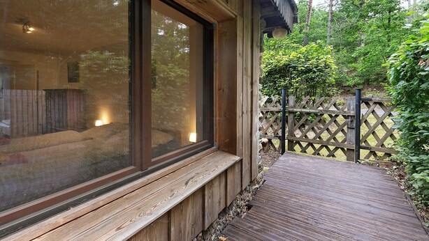 A wooden deck leading to a building with large windows and a lattice fence in the background. - Bungalow 'Urlaubsoase Am Bruchsee' with Lake View, Private Terrace and Private Garden (Templin)