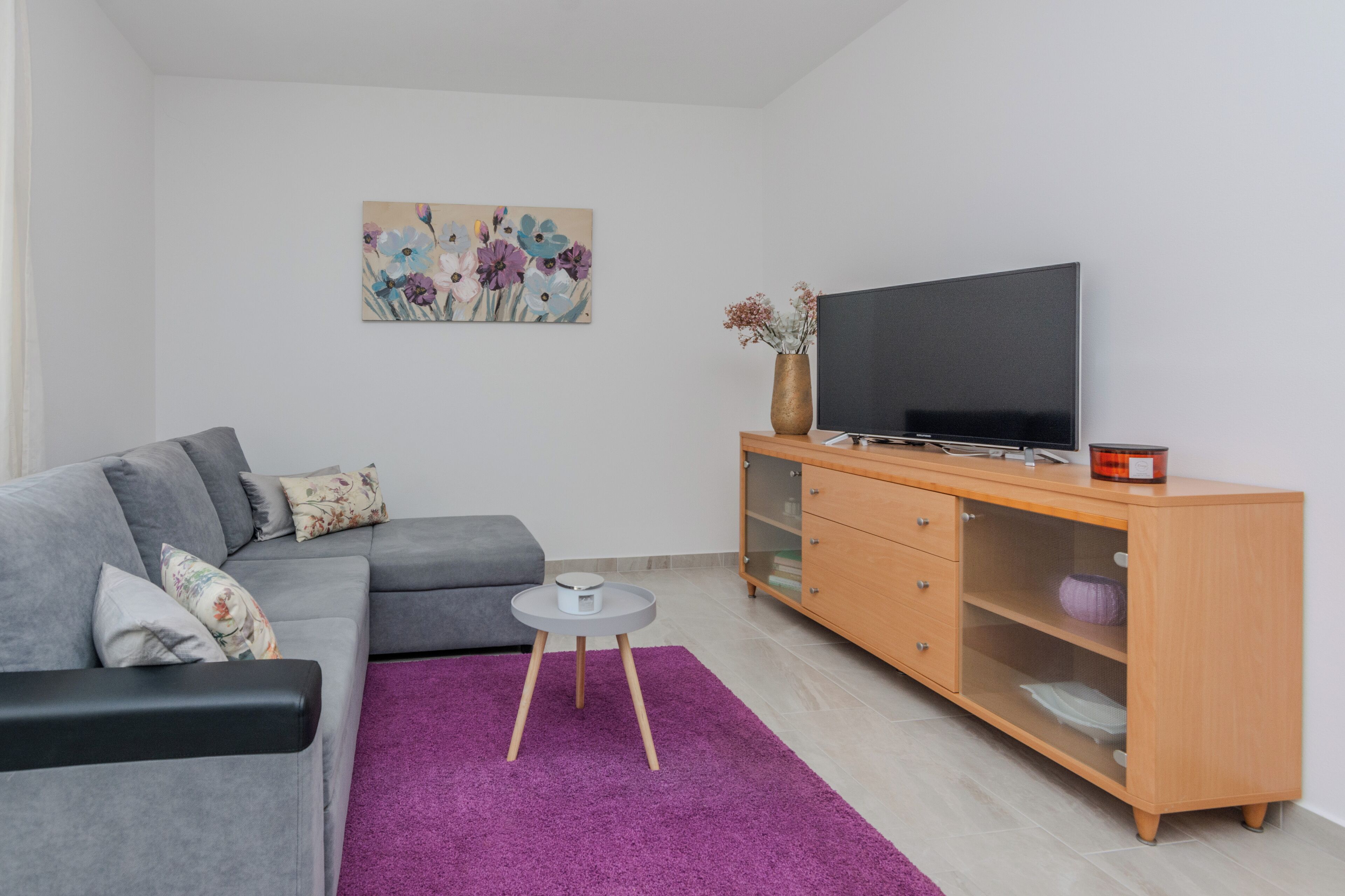 En moderne stue med en grå sofa, et rundt bord, et fjernsyn på et træskab og en lilla måtte.