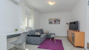 A modern living room with a grey sofa, a small round table, a purple rug, a TV on a wooden cabinet, and a window with curtains. - Holiday home Ella (Privlaka)
