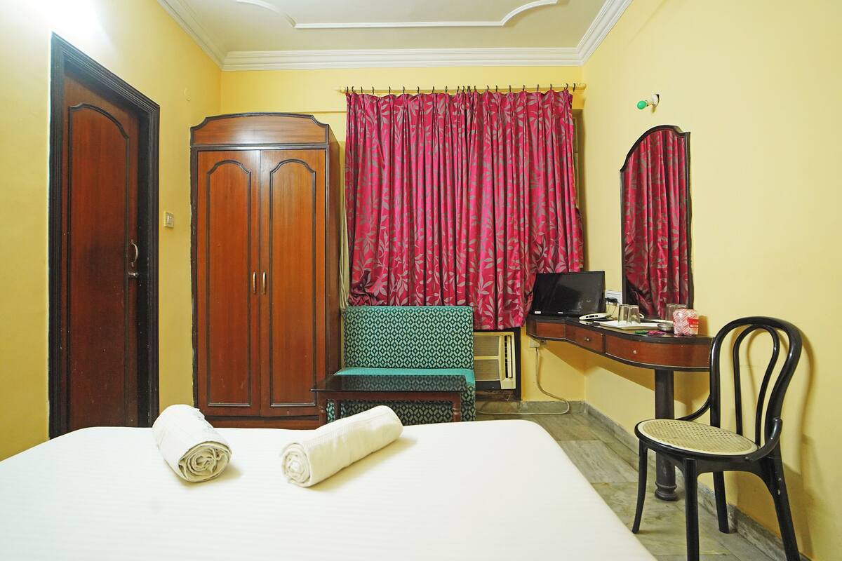 A hotel room with a bed, a wooden wardrobe, a desk with a chair, a television, and red curtains.