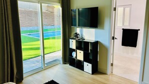 A modern living room with a large sliding glass door leading to a garden, a flat-screen TV mounted on a black entertainment unit, and a white door with a black towel hanging on it.