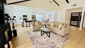A spacious living area with a sofa, coffee table, and a rug. There is a kitchen in the background with white cabinets and appliances.