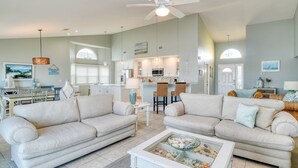 Ein geräumiges Wohnzimmer mit zwei beigen Sofas, einem Glastisch mit Meeresschnecken und einer dahinterliegenden Küche. Eine Deckenventilator ist ebenfalls zu sehen.