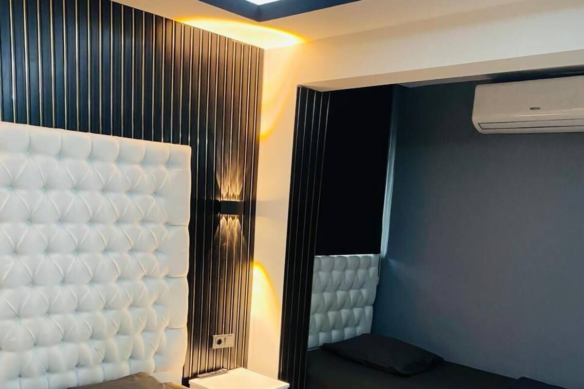 A modern bedroom with a tufted headboard, a bedside table, a bed with dark bedding, and an air conditioning unit.