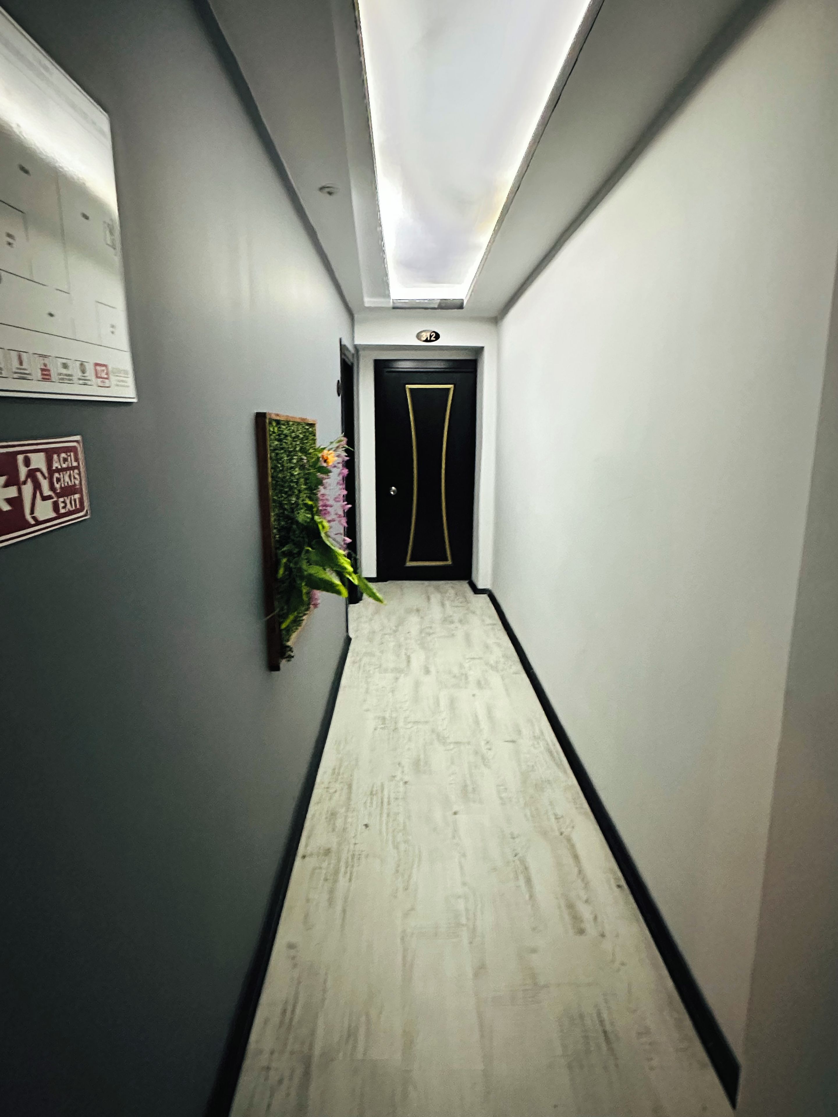 Un pasillo estrecho con una puerta al final, un letrero de salida de emergencia en la pared y una planta en maceta.