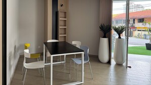 A modern dining area with a black table and white chairs, a wooden bookshelf, and large potted plants. - Havona Hotel @ CIQ Johor Bahru - Self Check-In (Johor Bahru)