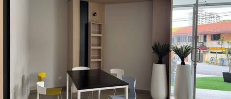 Un comedor moderno con una mesa negra y sillas blancas, una estantería de madera y plantas grandes en macetas.