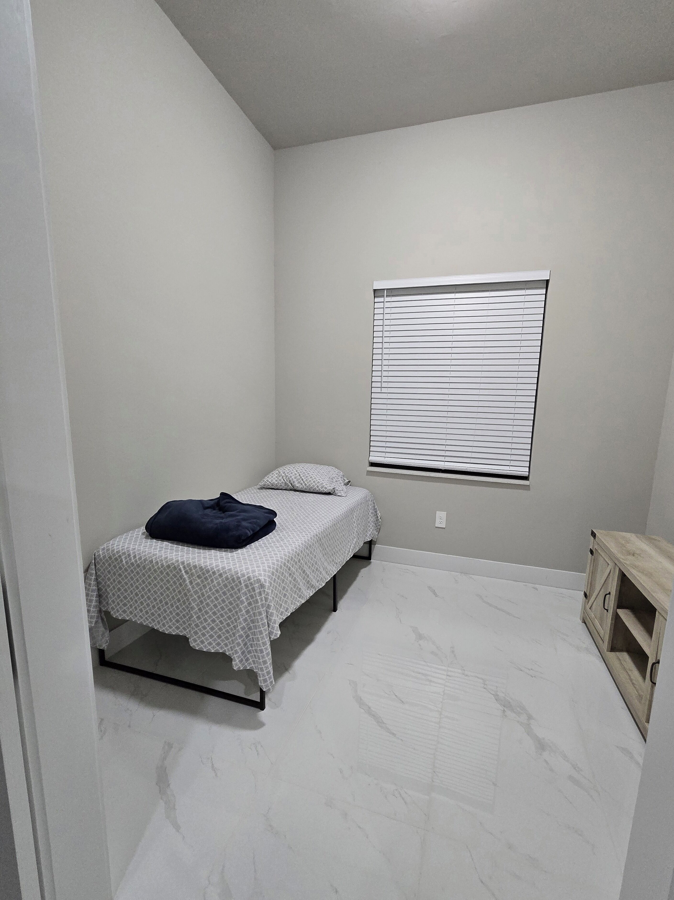 A small, sparsely furnished room with a bed, a window with blinds, and a small wooden cabinet.