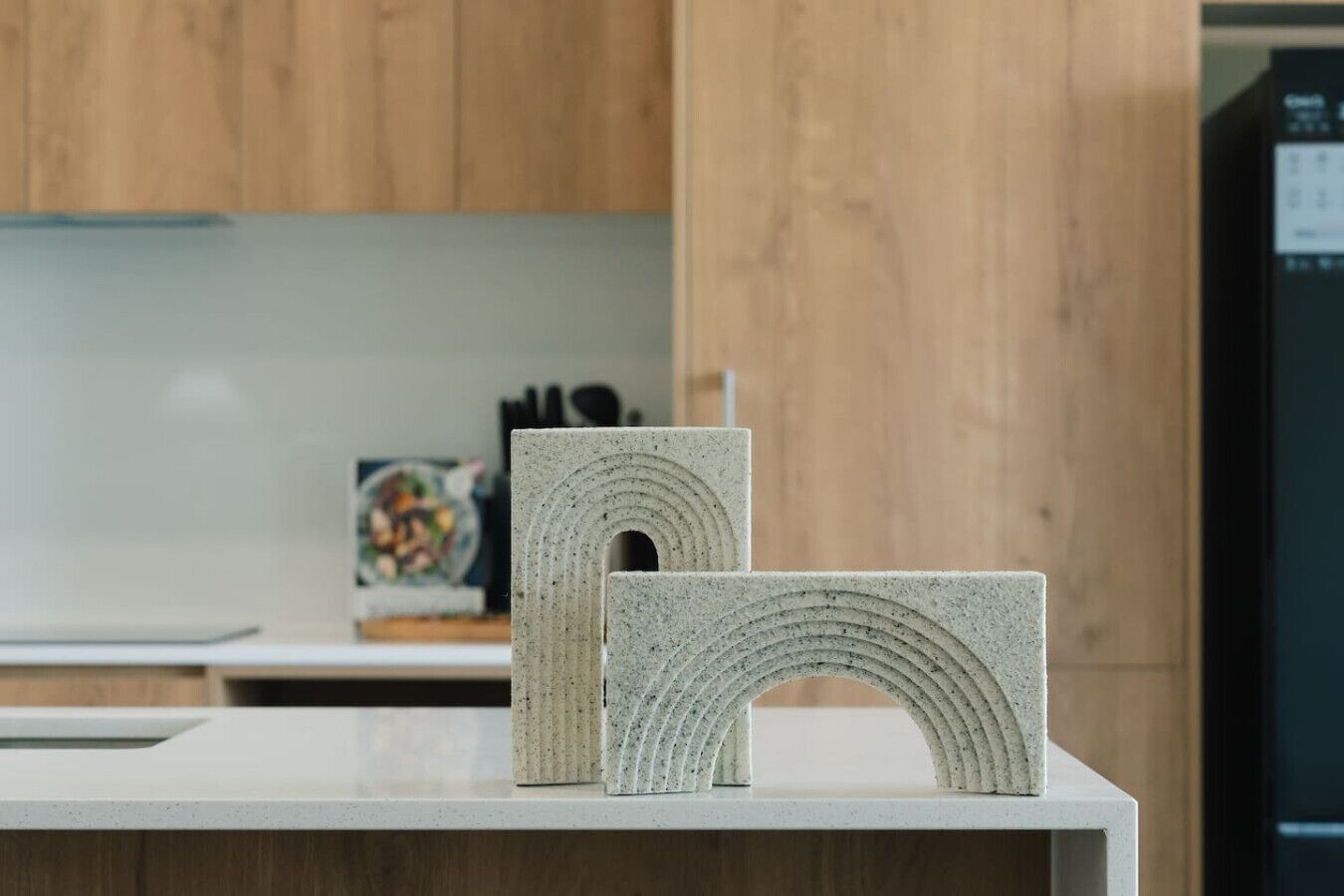 Two stone sculptures on a countertop in a kitchen with wooden cabinets and a refrigerator in the background.