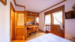 Una habitación con una cama, un escritorio de madera con sillas, una ventana con cortinas y un televisor montado en la pared.