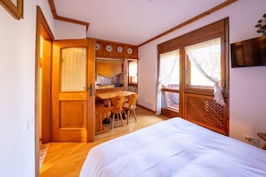 A room with a bed, a wooden desk with chairs, a window with curtains, and a TV mounted on the wall.