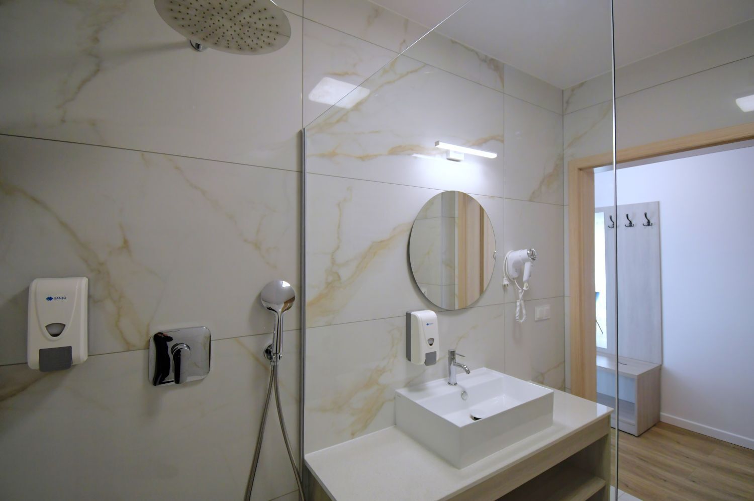 A modern bathroom with a marble backsplash, a round mirror, a handheld showerhead, a wall-mounted hairdryer, and a white sink.